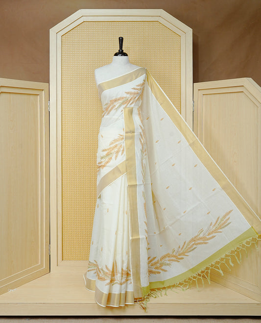 Cream Colour Cotton Kerala Saree Featuring Delicate Wheat Motifs and Scattered Leaf Designs On Body, Enhanced with a Classic Gold Zari Border. The Pallu Is Finished with Light Green Tassels and Paired with a Matching Blouse Piece.