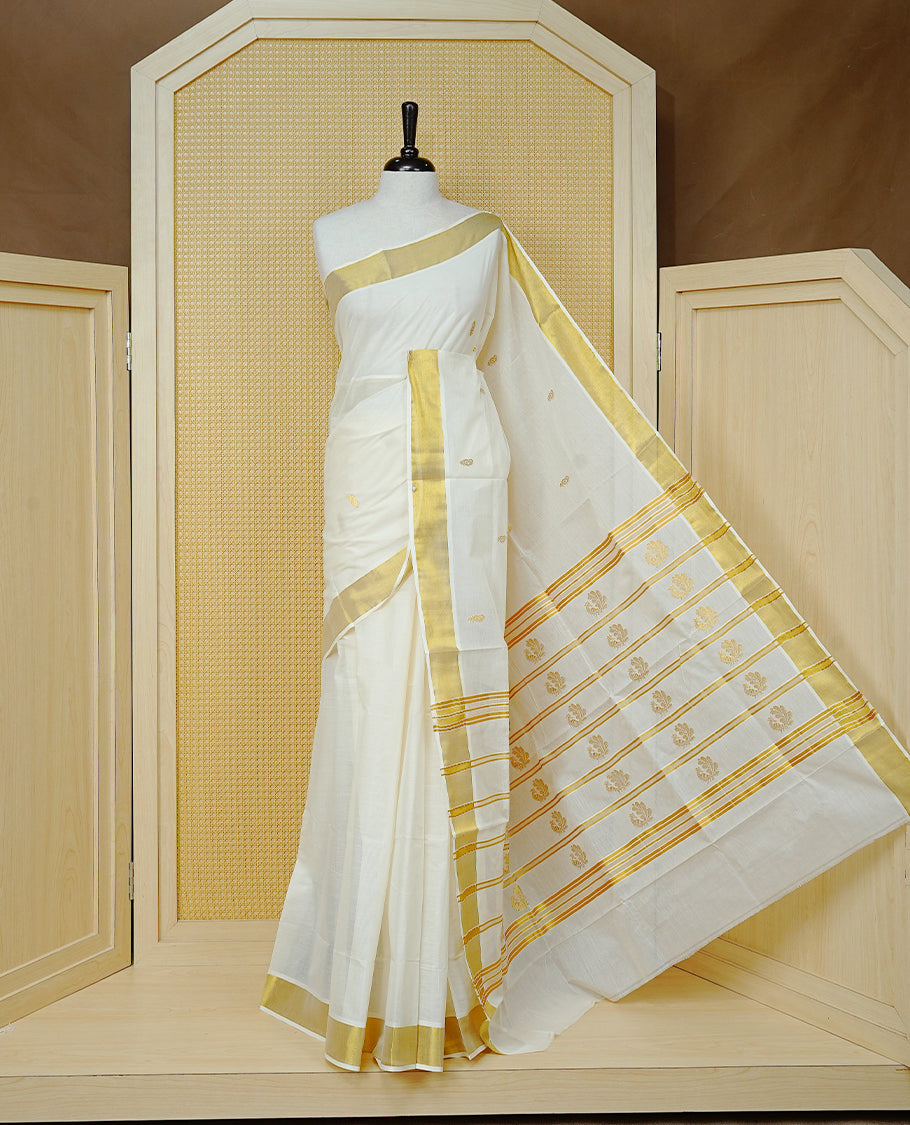 Cream Colour Cotton Kerala Saree Featuring Delicate Gold Leaf  Pattern Across The Body, Enhanced With A Traditional Gold Zari Border, A Gold Peacock Motifs &  Striped Gold Zari Pallu, Finished With A Matching Blouse Piece