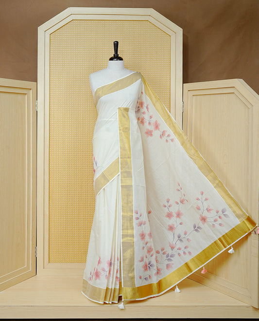 Cream Colour  Cotton Kerala Saree Featuring Digital Floral Prints Across The Body, Enhanced With A Gold Zari Border, Traditional Gold Zari Pallu, Finished With Peach & White  Tassels And A Matching Blouse Piece.