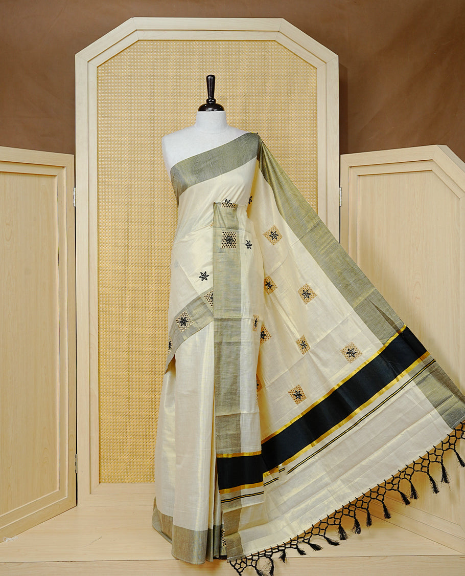 Cream Colour Tissue Kerala Saree Featuring Delicate Black Floral Embroidery Work Across The Body, Enhanced With A Grey Zari Border And A Rich  Black & Gold Zari Pallu, Finished With Black Tassels And A Matching Blouse Piece