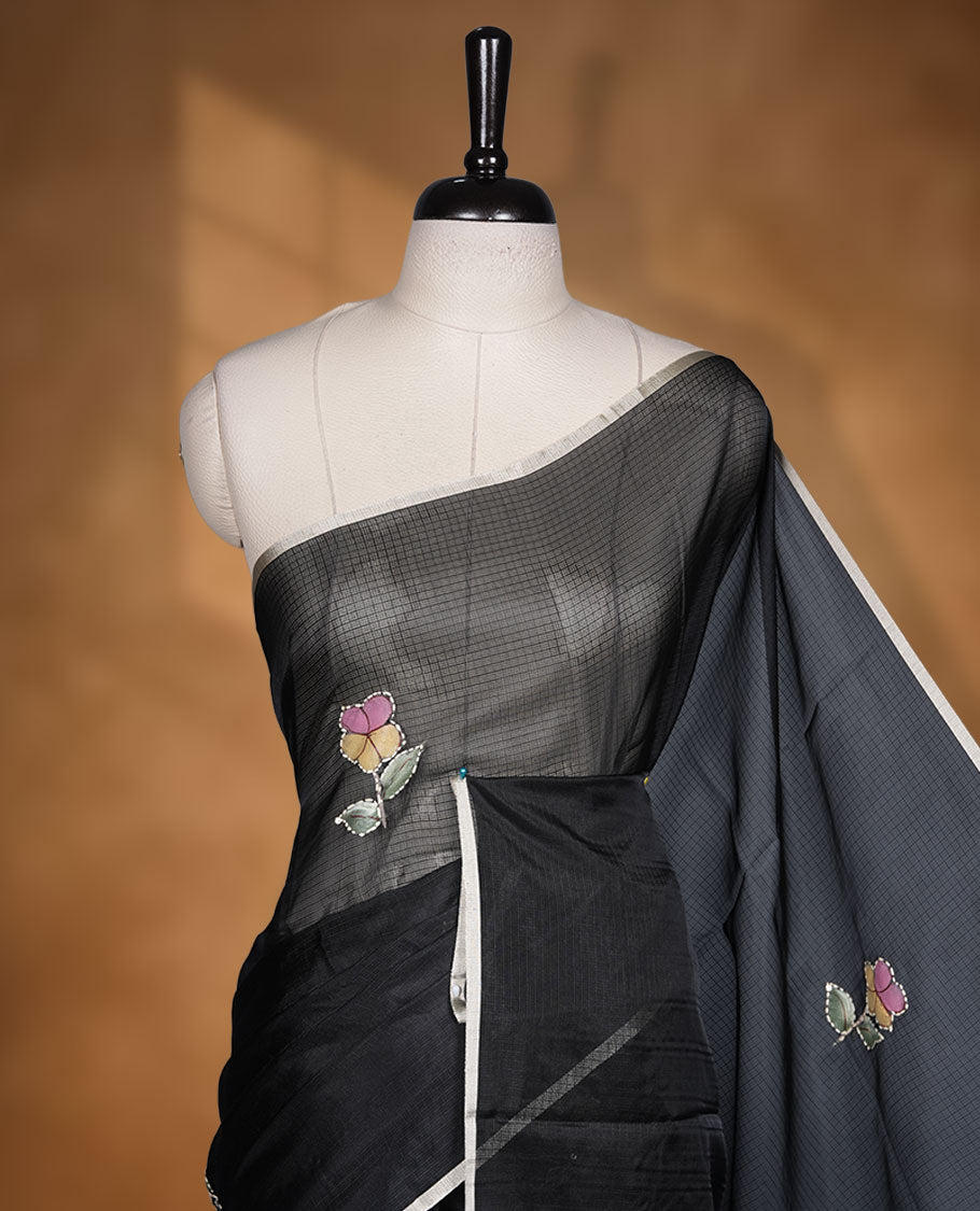 Black colour semi tusser silk saree showcasing delicate embroidered floral motifs throughout the body, finished with soft contrast detailing on the border and complemented by a matching embroidered blouse.