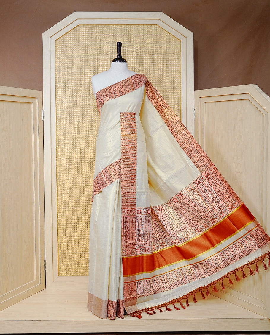 Cream Colour Tissue Kerala Saree Featuring a Striking Rust Red Printed Border with Traditional Geometric Motifs. The Rich Rust Pallu Is Accentuated with Gold Zari Detailing and Finished with Matching Tassels, Paired with a Matching Blouse Piece.