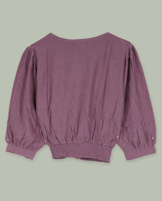 Dusty mauve three-quarter sleeve top with round neckline, metal button detail at the neck, and a pleated waistband accented with slim metallic trims.