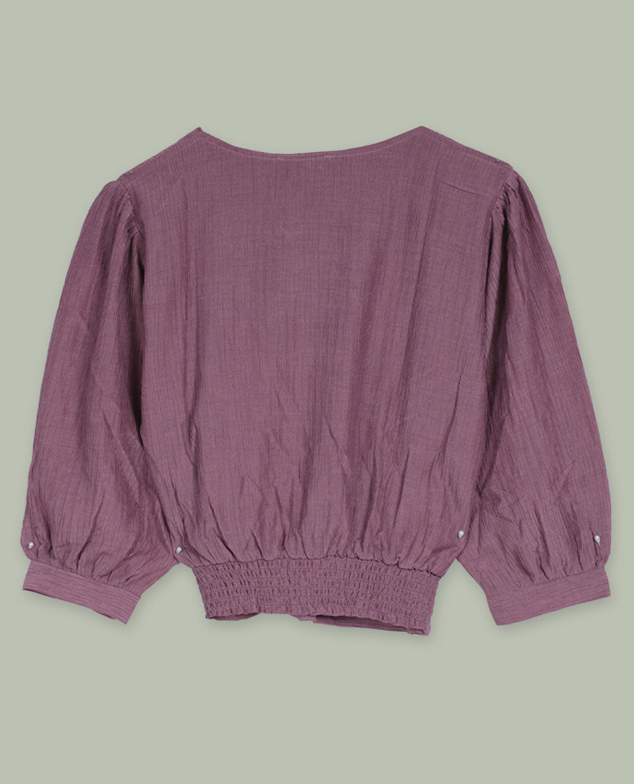 Dusty mauve three-quarter sleeve top with round neckline, metal button detail at the neck, and a pleated waistband accented with slim metallic trims.