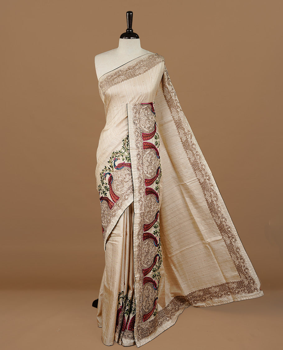 Cream colour Semi Tussar silk saree features an intricate cutwork embroidered peacock motifs on border, paired with a matching unstitched blouse piece.