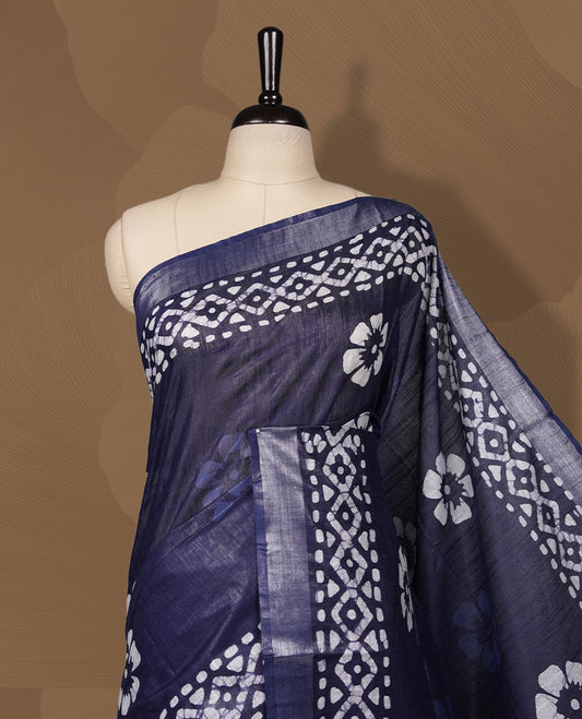 Navy blue colour semi jute tissue saree featuring white floral prints across the body, adorned with intricate geometric woven borders and a graceful floral work pallu & Comes with a matching blouse piece.