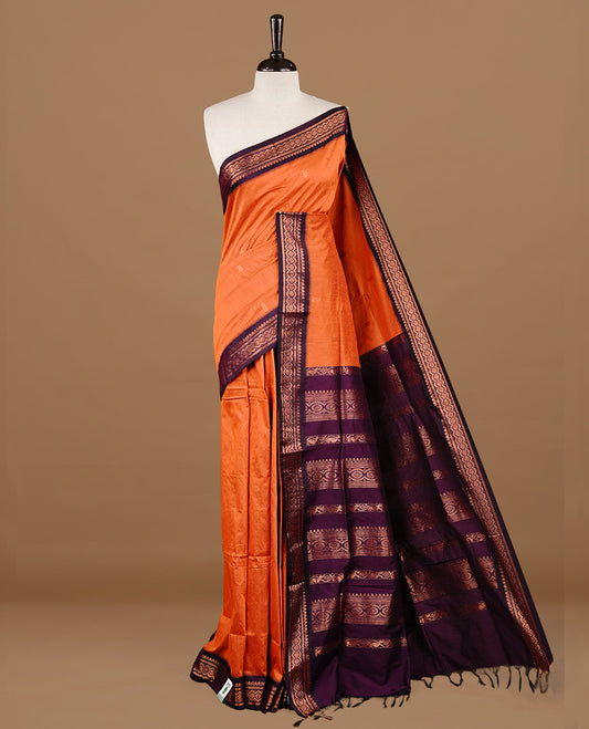 Orange colour kalyani cotton saree featuring gold zari motifs on body, highlighting contrast Purple colour with Antique gold zari border, paired with matching blouse piece.