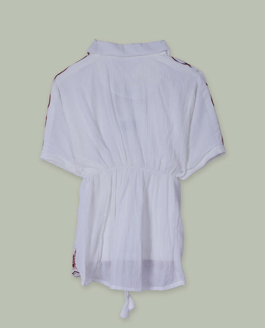 White short tunic top with collared neckline, short sleeves, contrast paisley and floral embroidery on shoulders and hem, and a drawstring waist with tassel ties.