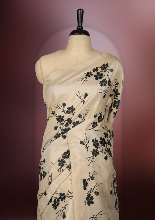 Beige colour saree adorned with black floral prints across the body, complemented by a striking black pallu with floral and stripe detailing. Finished with a subtle border and paired with a matching blouse.