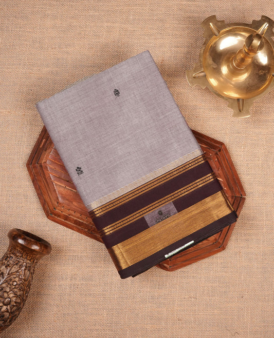 Grey colour chettinad cotton saree featuring small black buttas on body highlighting contrast dark chocolate colour with gold zari traditional border, pallu and paired with matching blouse piece.