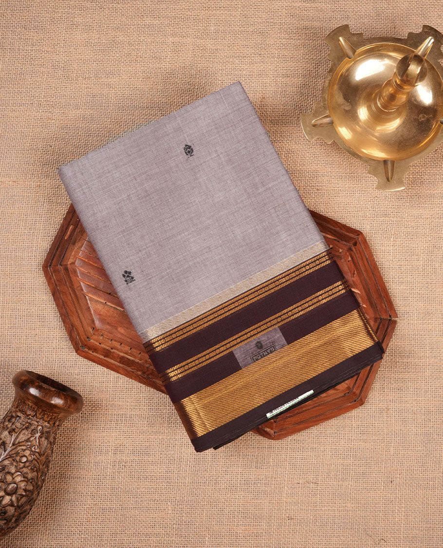 Grey colour chettinad cotton saree featuring small black buttas on body highlighting contrast dark chocolate colour with gold zari traditional border, pallu and paired with matching blouse piece.
