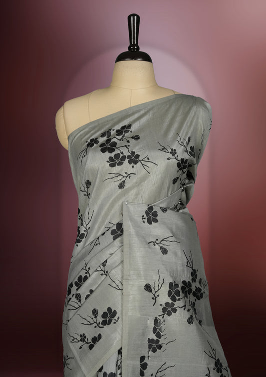 Grey colour saree with black floral prints all over the body, complemented by a contrasting black pallu adorned with bold floral designs and striped detailing & Paired with a matching blouse for a complete look.