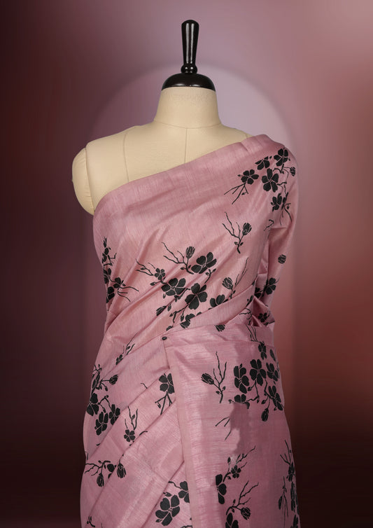 Pastel pink colour saree with black floral prints all over the body, complemented by a contrasting black pallu adorned with bold floral designs and striped detailing & Paired with a matching blouse for a complete look.