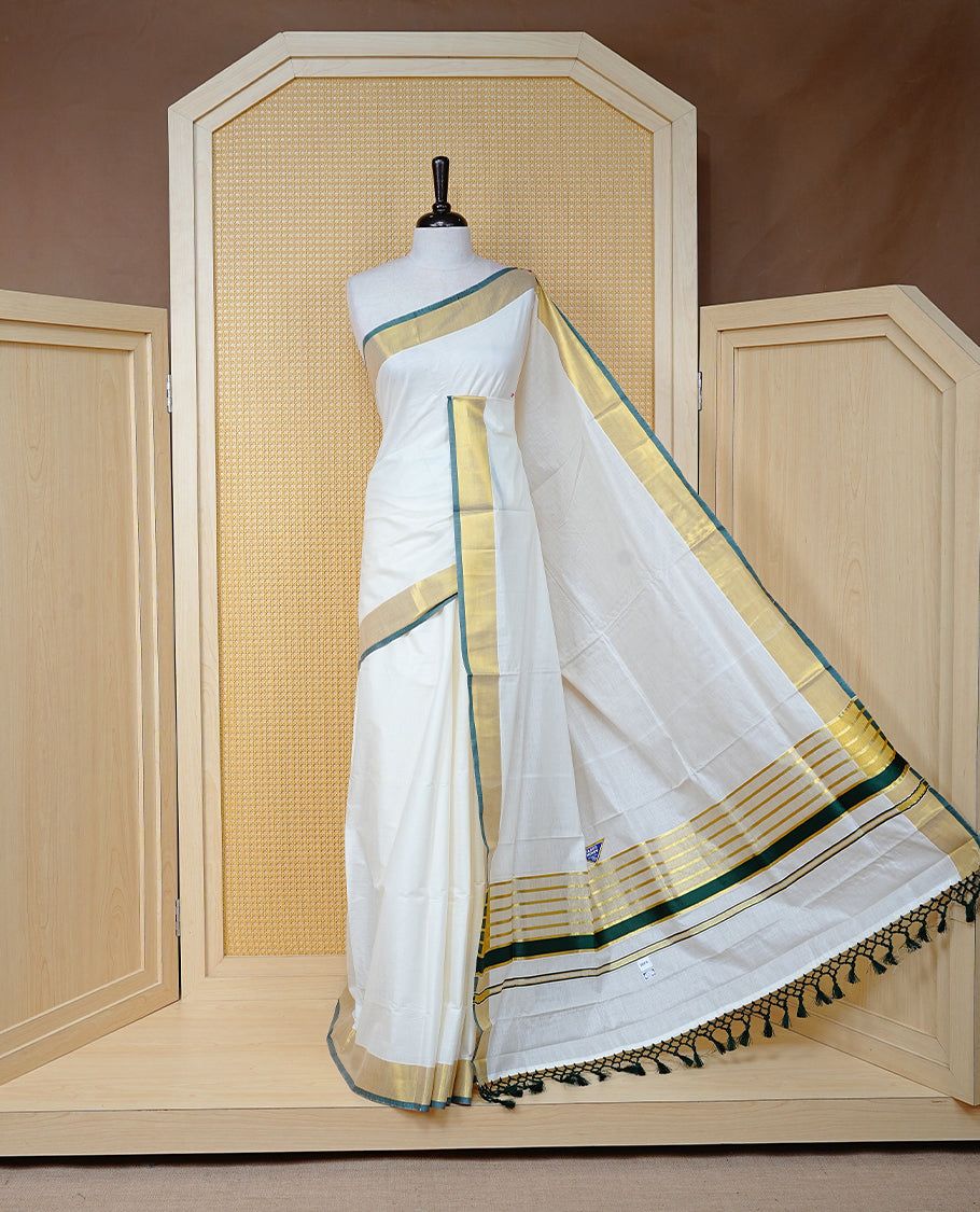 Cream Colour Cotton Kerala Saree Featuring A Plain Body, Enhanced With A Gold Zari & Green  Colour Border, A Traditional Gold Zari Pallu, Green Tassels, And A Matching Blouse Piece.