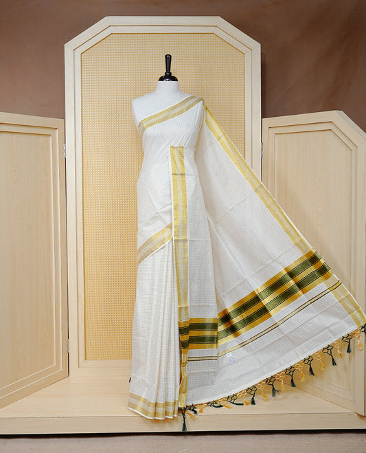 Cream Colour Cotton Kerala Saree Featuring A Golden Checks On Body, Enhanced With A Gold Zari  Border, A Traditional Gold Zari & Green Pallu, Green & Golden Tassels, And A Matching Blouse Piece.