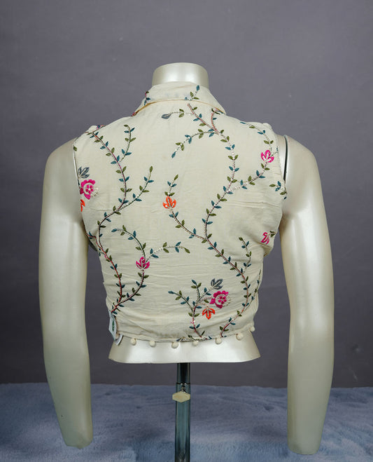 Cream colour sleeveless embroidered blouse with soft collar, front button detailing, delicate vine motifs, and pom-pom hem finish.
