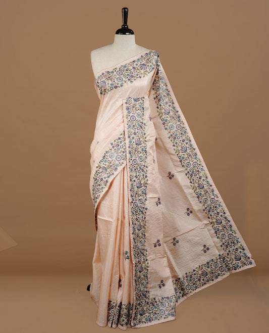 Pastel Peach colour Semi Tussar silk saree features an intricate, multi colored Kashmiri Kashida style floral embroidered border and scattered motifs, paired with a matching unstitched blouse piece.