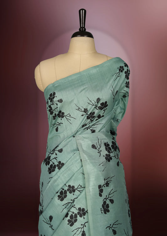Pastel green colour saree with black floral prints all over the body, complemented by a contrasting black pallu adorned with bold floral designs and striped detailing & Paired with a matching blouse for a complete look.