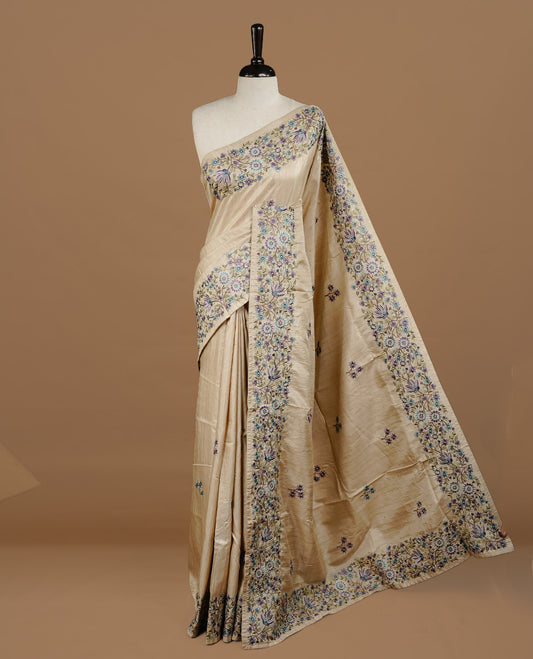 Cream grey colour Semi Tussar silk saree features an intricate, multi colored Kashmiri Kashida style floral embroidered border and scattered motifs, paired with a matching unstitched blouse piece.