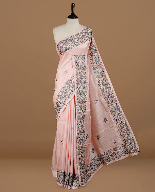 Pastel pink colour Semi Tussar silk saree features an intricate, multi colored Kashmiri Kashida style floral embroidered border and scattered motifs, paired with a matching unstitched blouse piece.