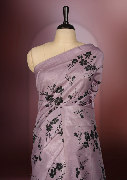 Pastel purple colour saree with delicate black floral prints spread across the body, The black pallu features coordinated floral motifs and stripes, Finished with a sleek border and paired with a matching blouse.