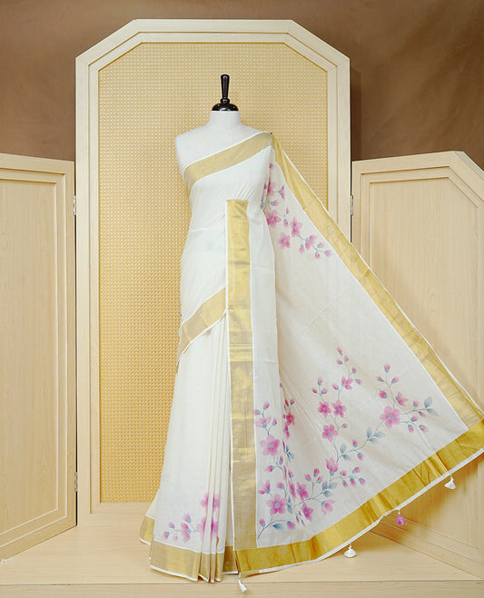 Cream Colour Cotton Kerala Saree Featuring Digital Pink Floral Prints Across The Body, Enhanced With A Gold Zari Border, Traditional Gold Zari Pallu, Finished With Pink & White Tassels And A Matching Blouse Piece