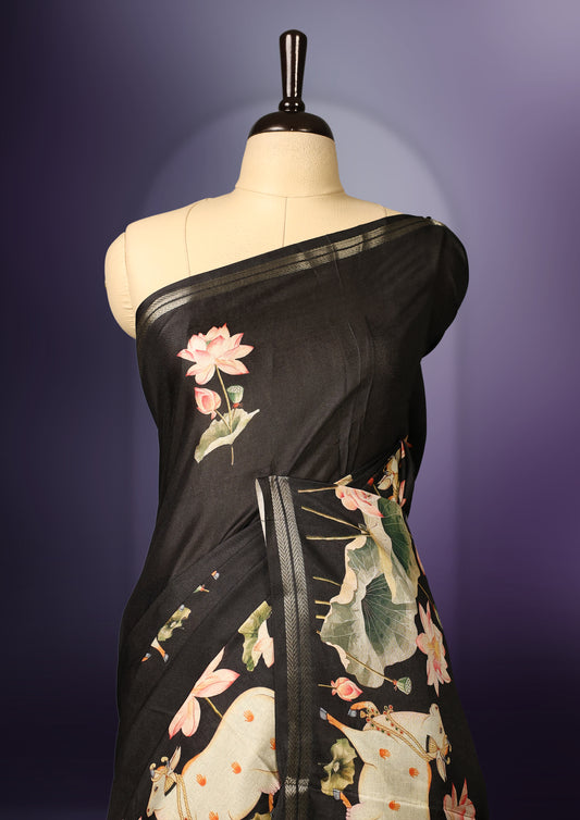 Black colour saree adorned with elegant lotus floral prints across the body and pallu, complemented by a sleek matching border, and paired with a coordinating blouse.