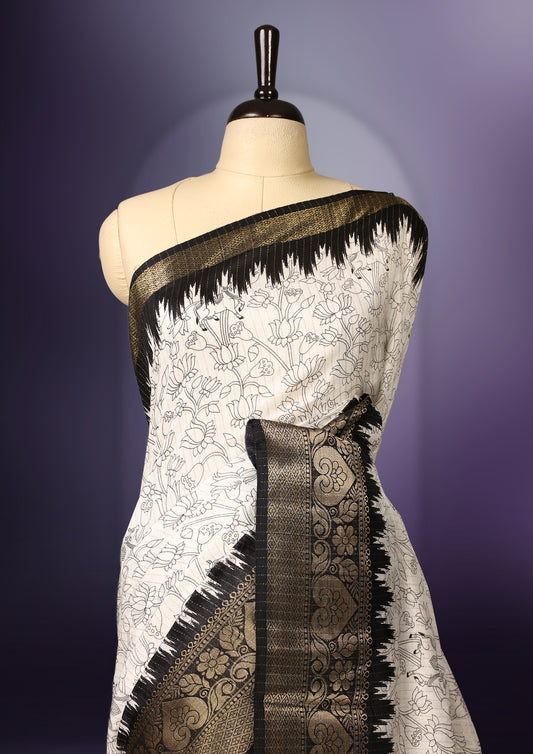 White colour saree adorned with intricate floral patterns across the body, enhanced by a rich black and gold zari border, featuring a vibrant artistic pallu with tree and nature motifs, paired with a matching blouse.