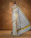 Off white colour cotton Kerala saree featuring colourful geometric motifs, complemented by a classic golden zari border and traditional pallu, paired with a matching blouse.