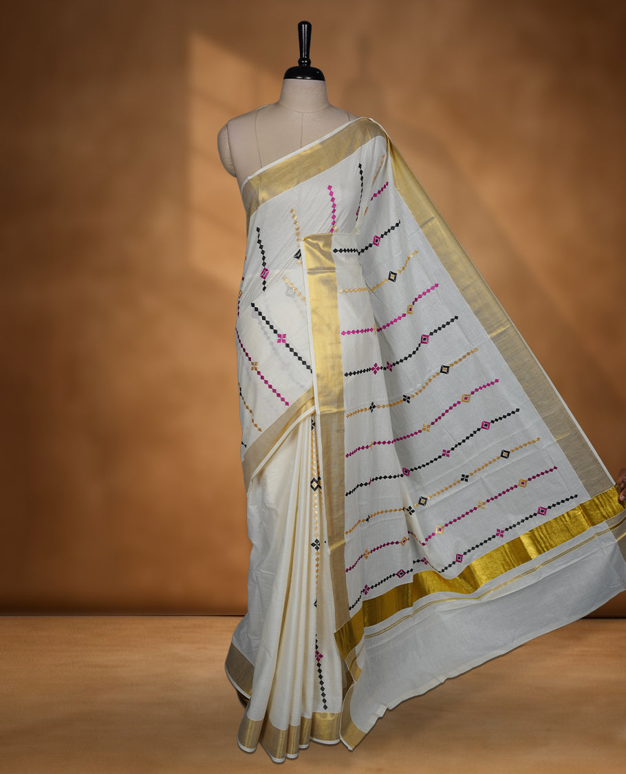 Off white colour cotton Kerala saree featuring colourful geometric motifs, complemented by a classic golden zari border and traditional pallu, paired with a matching blouse.