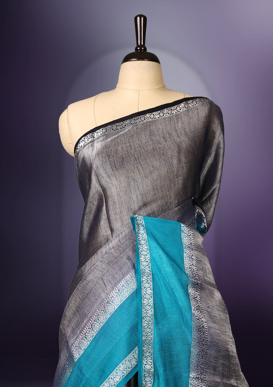 Steel grey saree with a rich sheen, paired with a contrasting turquoise blue border and pallu featuring delicate silver zari work, finished with tassels at the edge. Comes with a matching blouse piece.