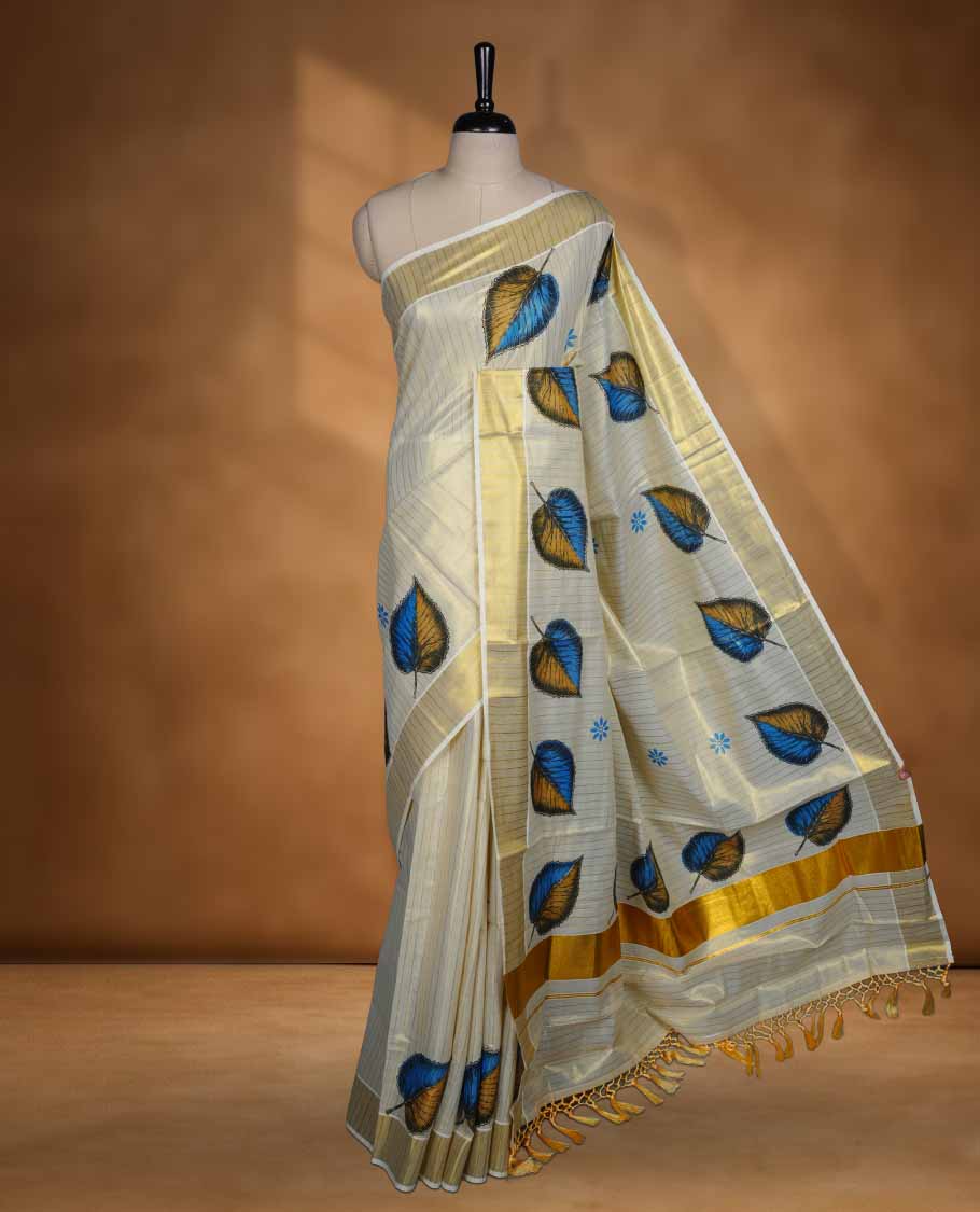 Off white colour Tissue Kerala saree featuring a subtle striped body with elegant blue and mustard leaf prints, complemented by a classic golden zari border and tasselled pallu, paired with a matching blouse.