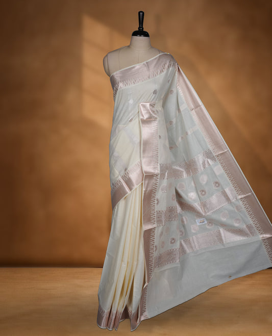 Off white colour cotton Kerala saree featuring subtle woven motifs, complemented by a soft rose gold zari border and rich traditional pallu, paired with a matching blouse.