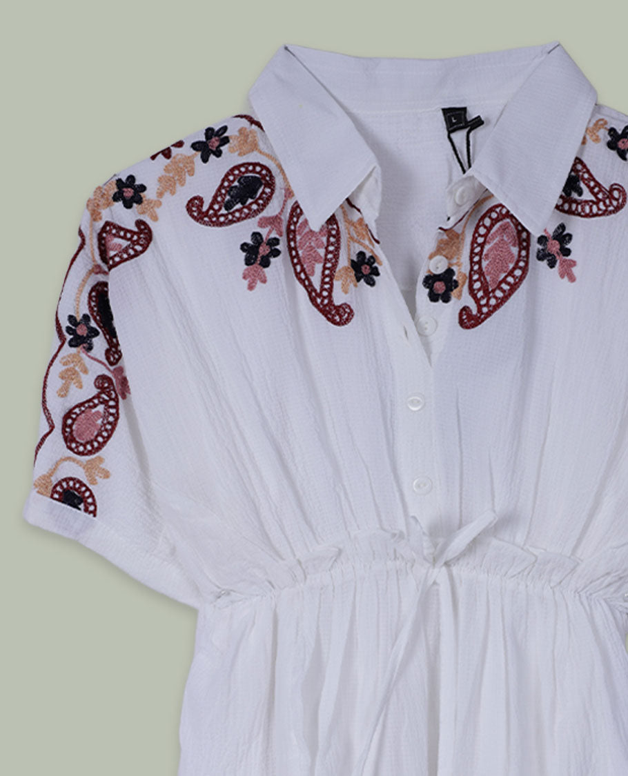 White short tunic top with collared neckline, short sleeves, contrast paisley and floral embroidery on shoulders and hem, and a drawstring waist with tassel ties.