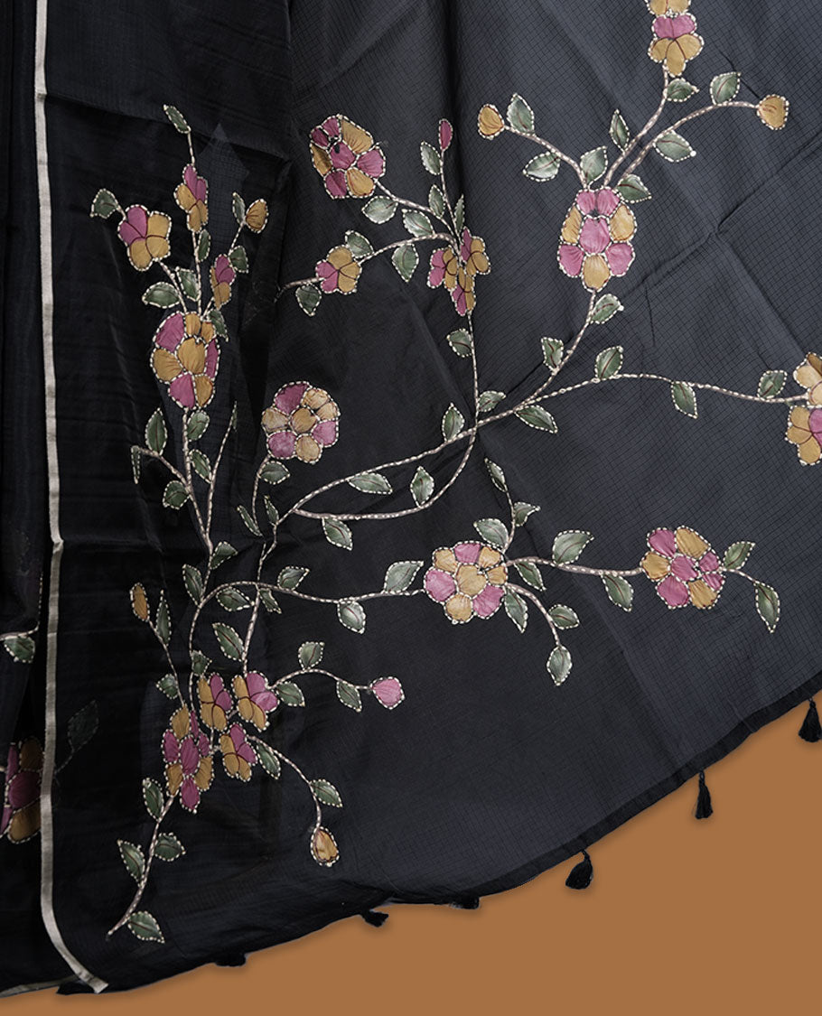Black colour semi tusser silk saree showcasing delicate embroidered floral motifs throughout the body, finished with soft contrast detailing on the border and complemented by a matching embroidered blouse.