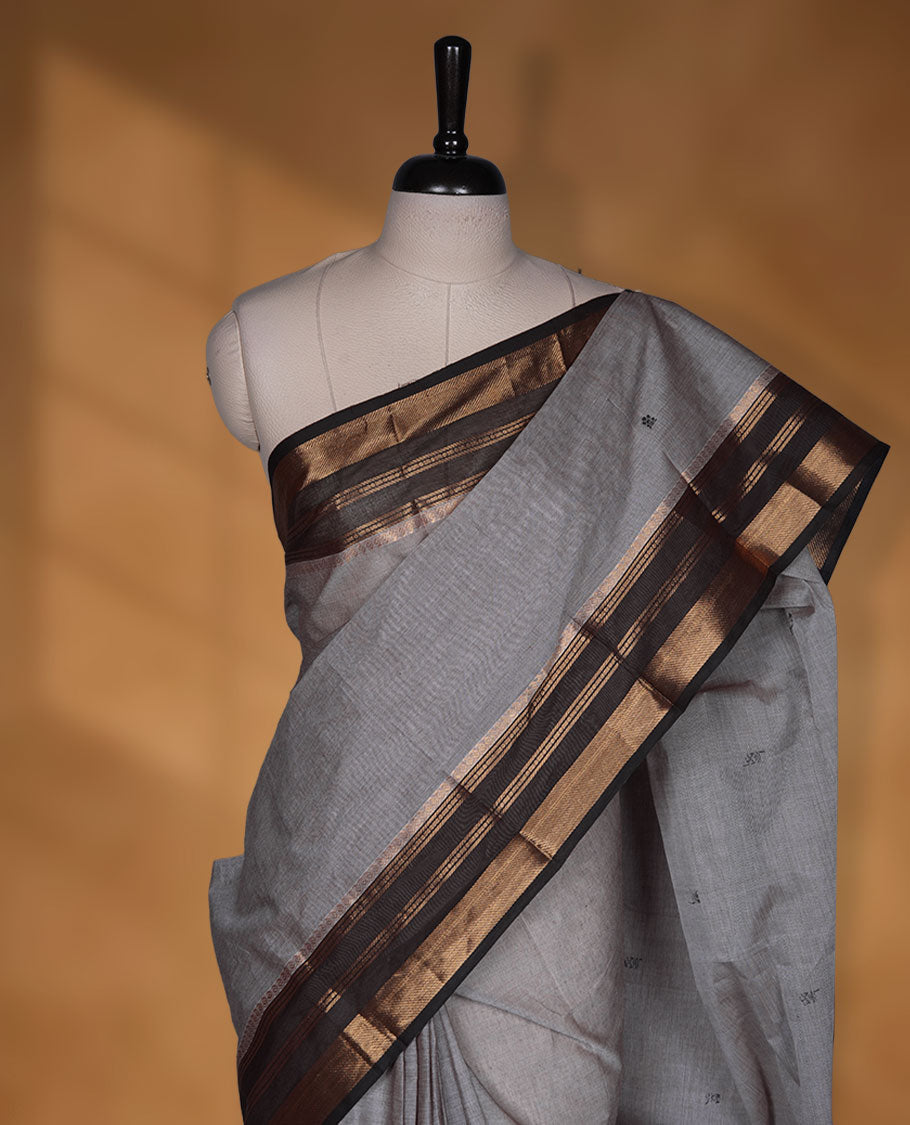 Grey colour chettinad cotton saree featuring small black buttas on body highlighting contrast dark chocolate colour with gold zari traditional border, pallu and paired with matching blouse piece.