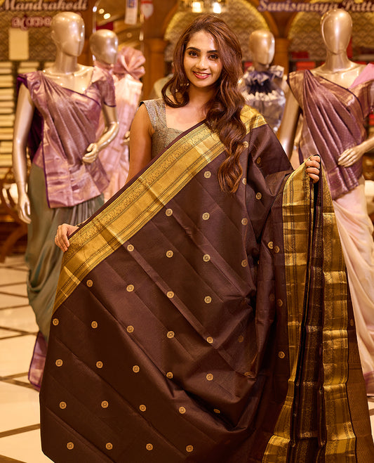 Graceful deep chocolate brown colour Kancheepuram silk saree adorned with delicate gold zari motifs across the body, beautifully finished with a broad traditional gold zari border, exuding timeless elegance and paired with a matching blouse piece