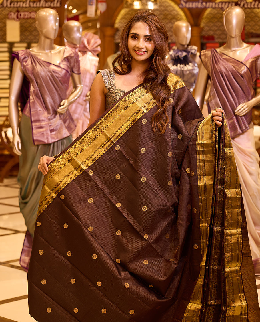 Graceful deep chocolate brown colour Kancheepuram silk saree adorned with delicate gold zari motifs across the body, beautifully finished with a broad traditional gold zari border, exuding timeless elegance and paired with a matching blouse piece