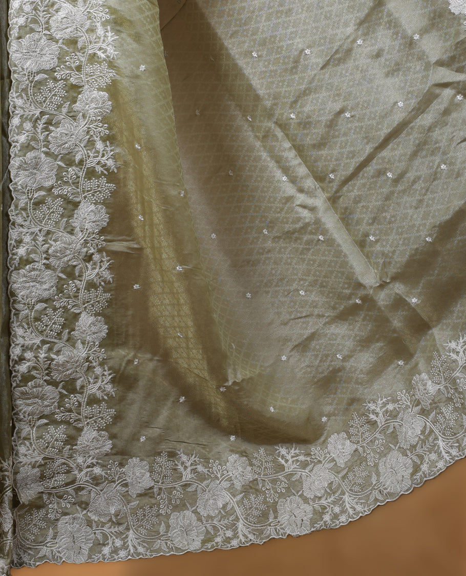 Pastel olive colour organza designer saree featuring a subtle self patterned body and an intricately scalloped floral thread embroidered border all around, complemented by a coordinating embroidered pallu and paired with a matching blouse.