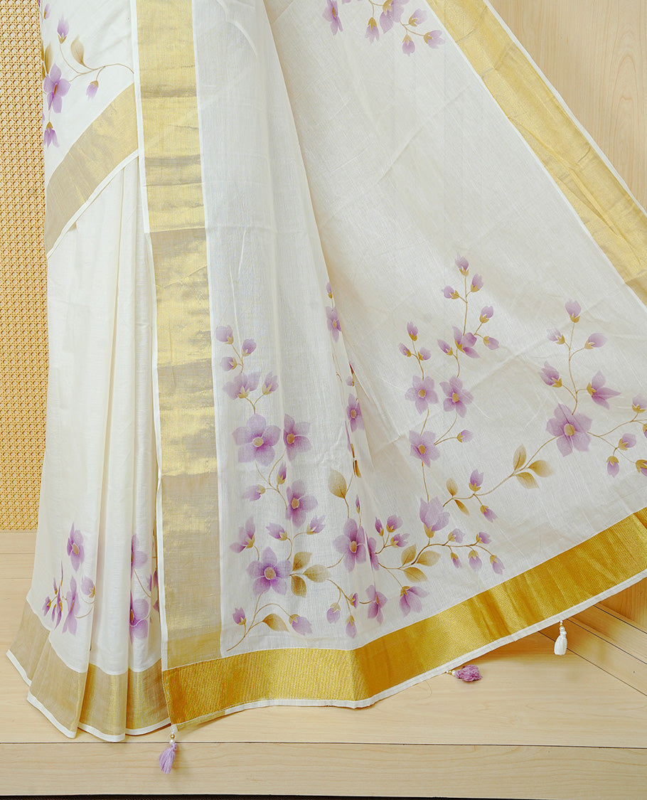 Cream Colour Cotton Kerala Saree Featuring Digital  Purple Floral Prints Across The Body, Enhanced With A Gold Zari Border, Traditional Gold Zari Pallu, Finished With Purple & White Tassels And A Matching Blouse Piece