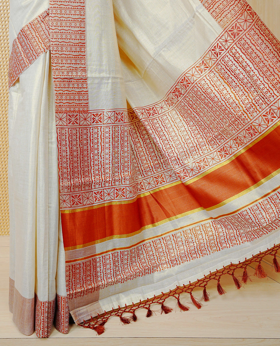 Cream Colour Tissue Kerala Saree Featuring a Striking Rust Red Printed Border with Traditional Geometric Motifs. The Rich Rust Pallu Is Accentuated with Gold Zari Detailing and Finished with Matching Tassels, Paired with a Matching Blouse Piece.