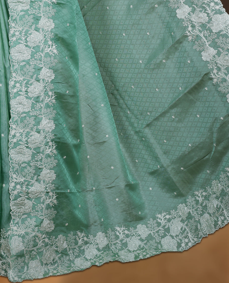 Light Green colour organza designer saree featuring a subtle self patterned body and an intricately scalloped floral thread embroidered border all around, complemented by a coordinating embroidered pallu and paired with a matching blouse.