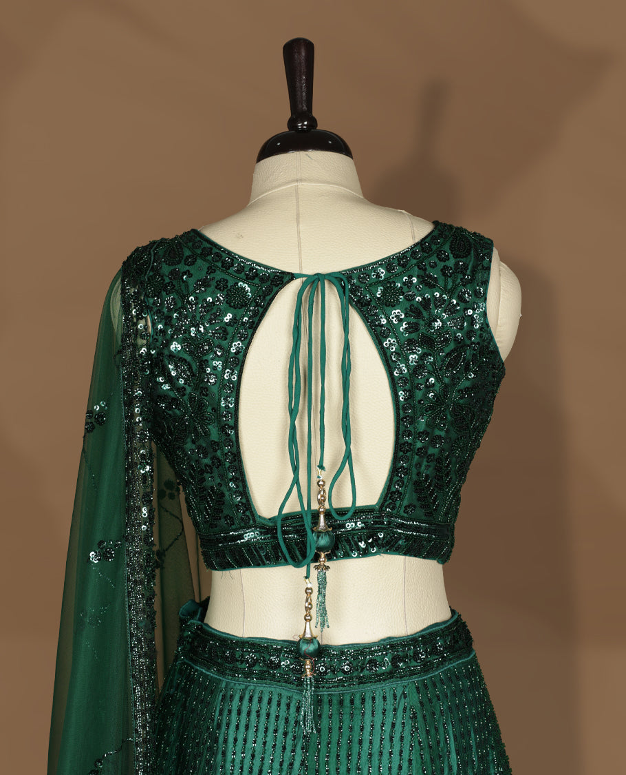 Emerald green colour lehenga set with stitched sleeveless V‑neck heavily sequinned blouse featuring cut‑out tie‑up back, matching stitched flared lehenga, and net dupatta with sequin border and scattered floral motifs.