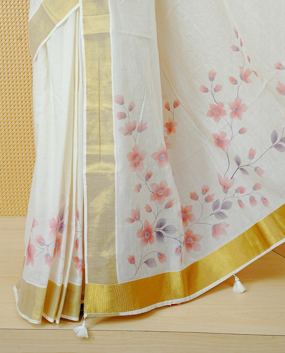 Cream Colour  Cotton Kerala Saree Featuring Digital Floral Prints Across The Body, Enhanced With A Gold Zari Border, Traditional Gold Zari Pallu, Finished With Peach & White  Tassels And A Matching Blouse Piece.