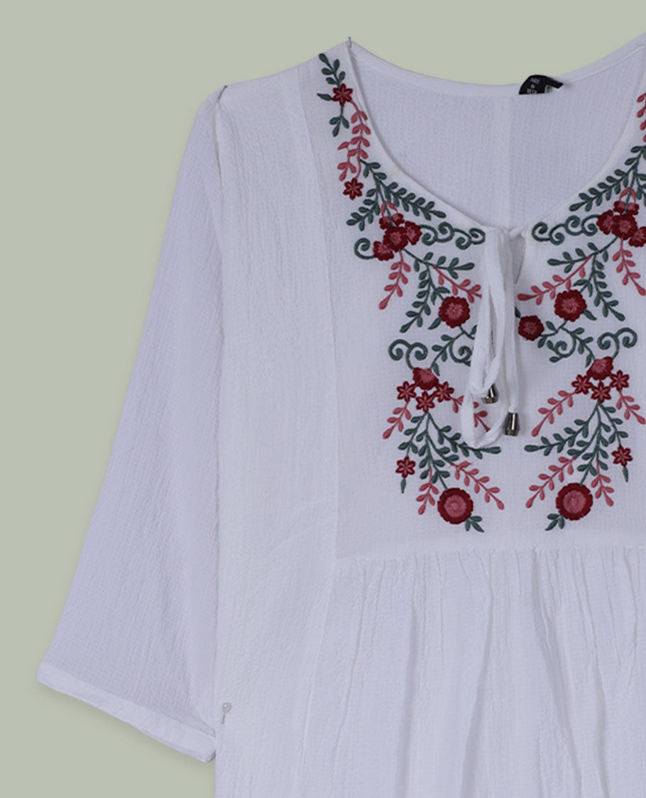 White three-quarter sleeve tunic top with round neckline and front yoke featuring red and green floral embroidery with tie-up detail at the neck.