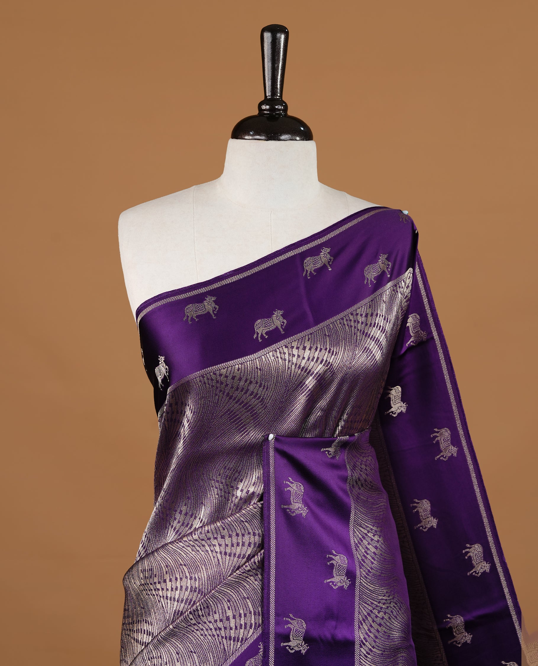 Purple colour semi Banaras crepe silk saree featuring an elegant gold zari patterned body, highlighted by a plain border adorned with traditional gold zari cow motifs. Paired with a matching blouse piece.