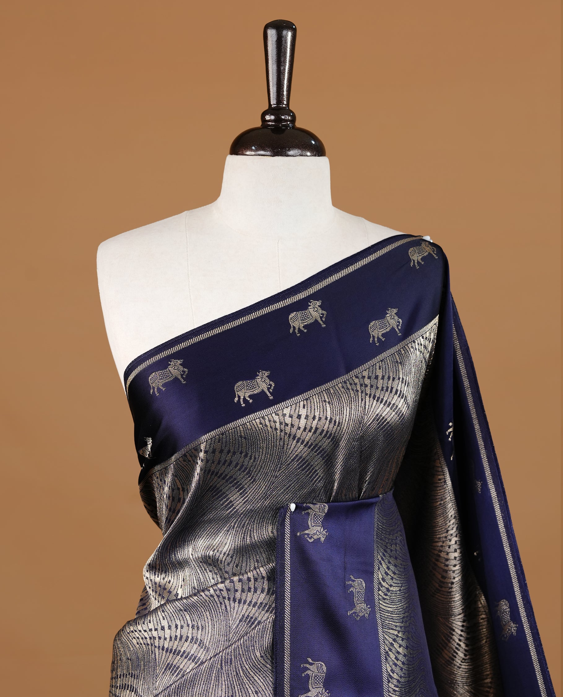 Dark Blue colour semi Banaras crepe silk saree featuring an elegant gold zari patterned body, highlighted by a plain border adorned with traditional gold zari cow motifs. Paired with a matching blouse piece.
