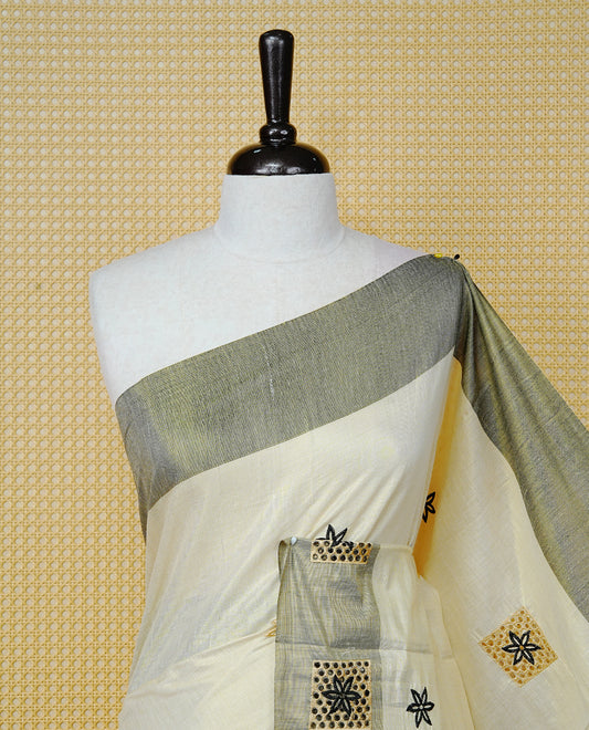 Cream Colour Tissue Kerala Saree Featuring Delicate Black Floral Embroidery Work Across The Body, Enhanced With A Grey Zari Border And A Rich  Black & Gold Zari Pallu, Finished With Black Tassels And A Matching Blouse Piece