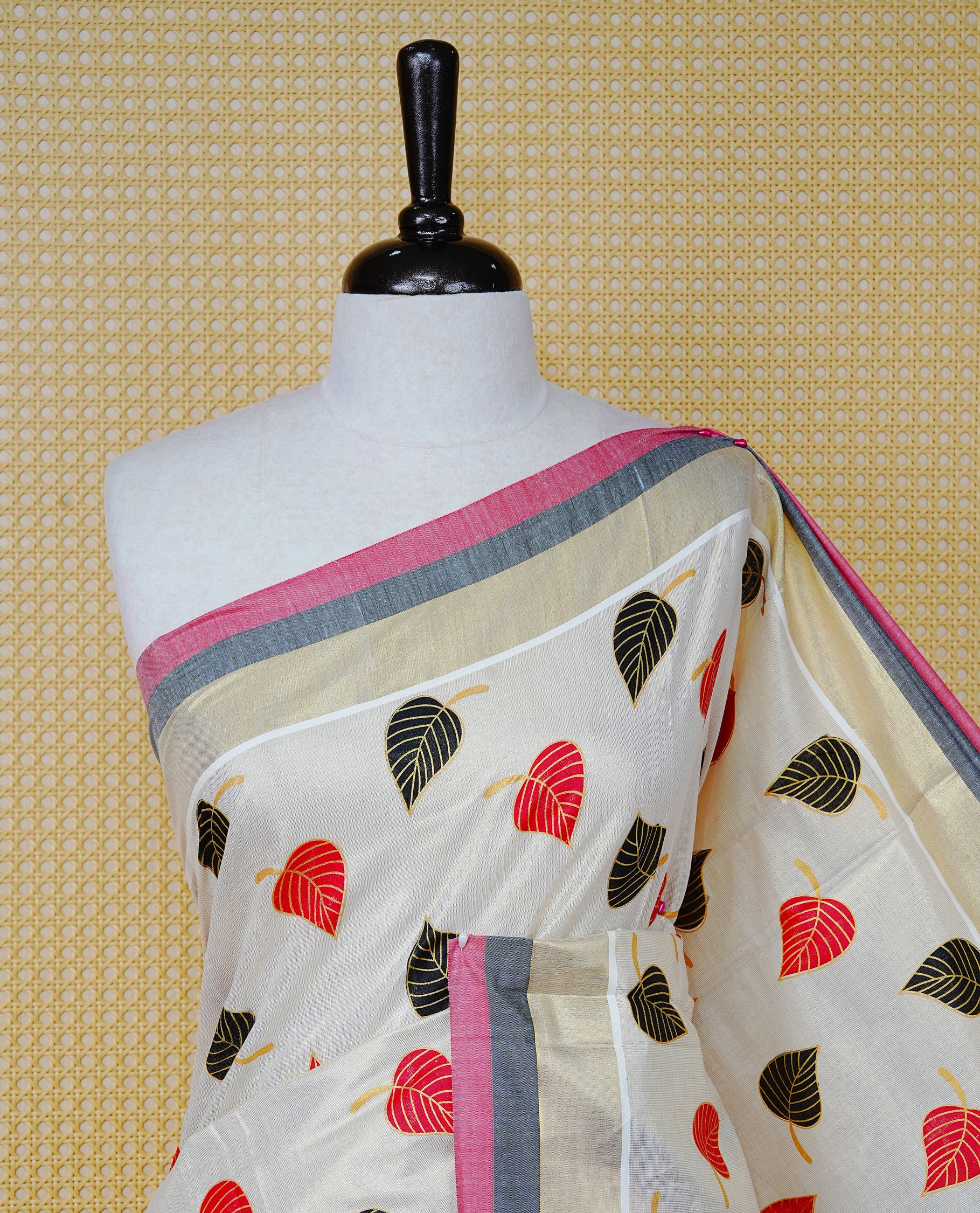 Cream Colour Tissue  Kerala Saree Featuring Red and Black Leaf Prints Across the Body, Enhanced with a Gold Zari, Red and Black Border &  Pallu, Red and Black Tassels, and a Matching Blouse Piece.