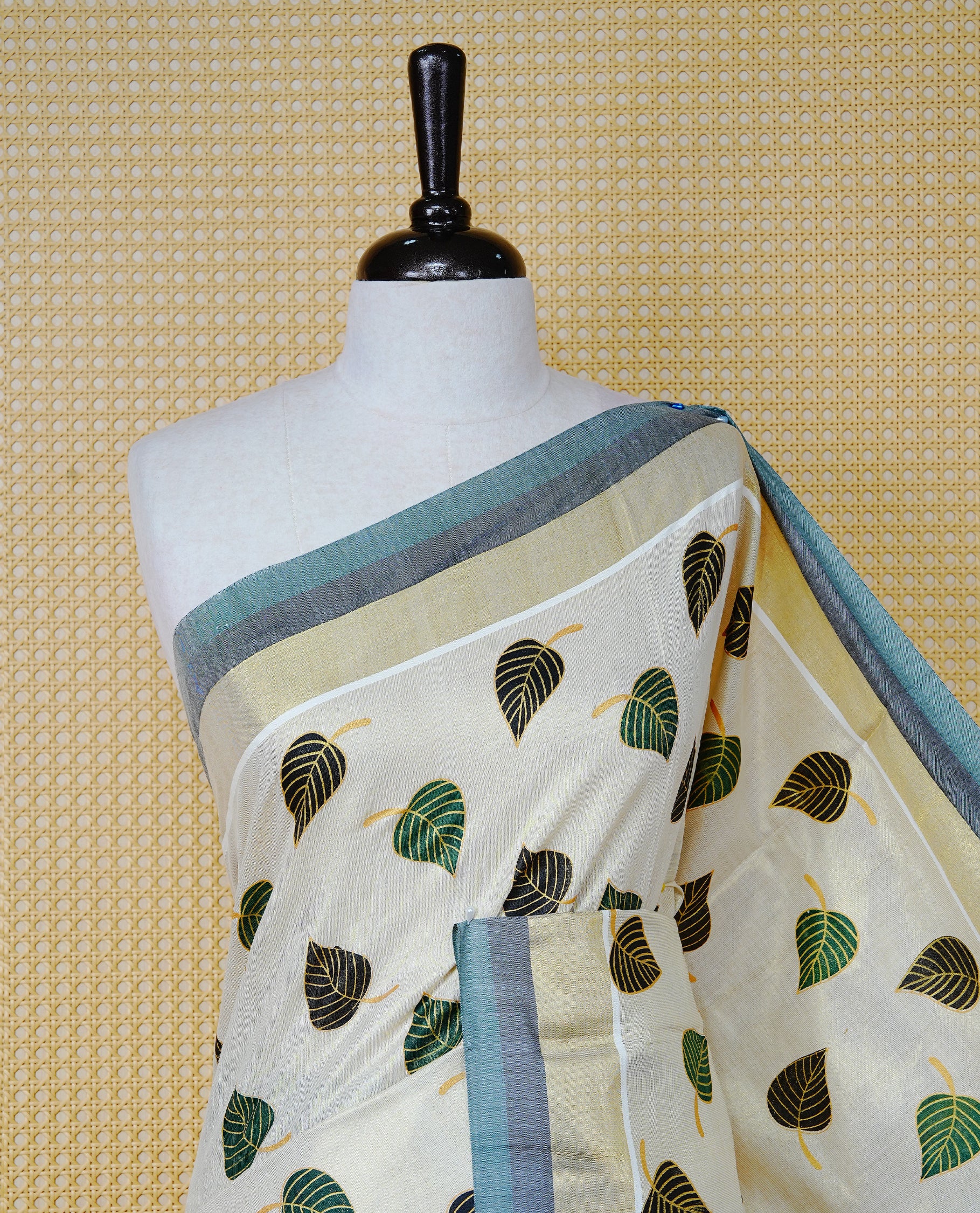 Cream Colour Tissue Kerala Saree Featuring Green and Black Leaf Prints Across the Body, Enhanced with a Gold Zari, Dark Green and Black Border & Pallu, Green  and Black Tassels, and a Matching Blouse Piece.